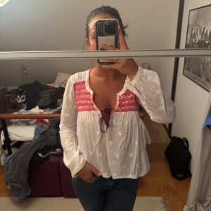 Vit blus med broderad rosa detalj - Supersöt vit blus med broderade blommor och en bred rosa broderad panel upptill. Blusen har v-ringning med knytband och långa ärmar. Luftig och flowy passform, perfekt för vår och sommar.