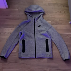 Grå Niketech hoodiejacka med dragkedja - Snygg grå hoodiejacka från Nike med svart logga på bröstet. Jackan har hel dragkedja framtill, två svarta fickor med dragkedja och en skön huva. Perfekt för chill eller träning, stilren och sportig look.
