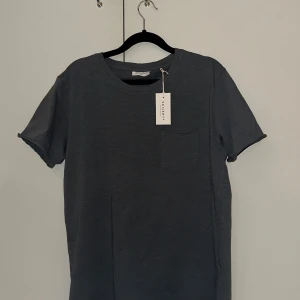 Mörkgrå t-shirt från Vailent - En stilren mörk grå/blå t-shirt från Vailent med rund halsringning och en liten bröstficka. T-shirten är i mjuk bomull och har klassisk passform med korta ärmar. Perfekt för en clean och enkel look. Den är också helt ny och aldrig använd.