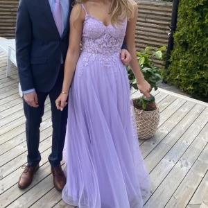 Ljuslila/syrenlila balklänning med spetsdetaljer - Säljer nu min balklänning som använts en gång! 💜💜 Det är en drömmig ljuslila långklänning med tunna axelband, v-ringning och blommiga spetsdetaljer upptill. Kjolen är i flera lager så att den inte blir genomskinlig, den har även en snygg slits framtill!🥰 Ingen storlek - kolla måtten på sista bilden. 