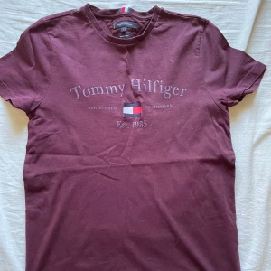 Vinröd t-shirt från Tommy Hilfiger - Snygg vinröd t-shirt från Tommy Hilfiger med klassisk logga och broderad flagga framtill. T-shirten har rund halsringning och korta ärmar. Tillverkad i mjuk bomull som känns skön mot huden. Perfekt för en chill och stilren look.