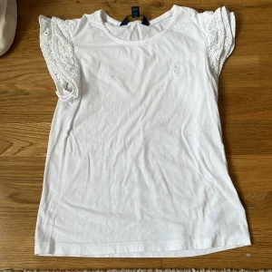 Vit blus från Polo Ralph Lauren - Supersöt vit blus från Polo Ralph Lauren med broderade, puffiga kortärmar i spets och klassisk rund halsringning. Liten broderad logga på bröstet. Perfekt för en fräsch och stilren look.