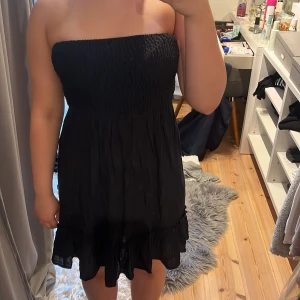 Svart axelbandslös kortklänning - Säljer en svart axelbandslös kortklänning med  volangkant nedtill. Klänningen har en luftig och lätt passform, perfekt för varma dagar. Materialet känns mjukt och bekvämt mot huden. Enkel och stilren design som är lätt att matcha.💖