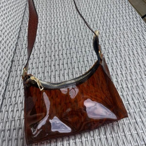 Brun transparent axelväska med dragkedja - Säljer en brun transparent axelväska i glansigt syntetmaterial. Väskan har en bred axelrem och stängs med en guldfärgad dragkedja. Perfekt för dig som gillar en unik och trendig look. Rymmer det viktigaste och är lätt att matcha med olika outfits.