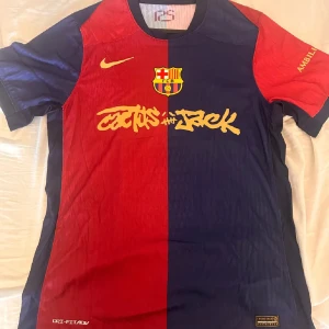 FC Barcelona x Cactus Jack Nike tröja - Säljer en FC Barcelona fotbollströja från Nike i samarbete med Cactus Jack. Tröjan har klassisk halva-halva design i rött och mörkblått, med gult tryck och klubbmärke på bröstet. Materialet är lätt och andas, perfekt för match eller träning.