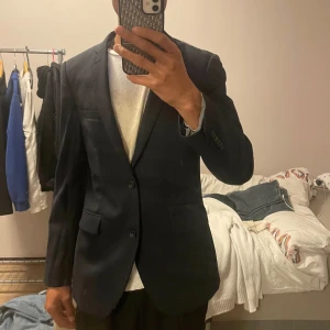 Mörkblå kavaj Riley Slim Fit - Snygg mörkblå kavaj från Riley i slim fit-modell. Klassisk design med två knappar framtill, fickor med lock och slät baksida. Fodrad insida i matchande blått tyg. Perfekt för dig som vill ha en stilren och modern look.