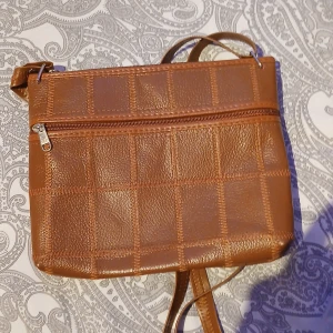 Brun rutmönstrad axelväska i skinn - Snygg brun axelväska med rutmönster och lång justerbar rem. Väskan är gjord i skinn och har en praktisk dragkedja upptill samt ett rymligt fack med röd insida. Perfekt för att bära med sig det viktigaste och samtidigt hålla stilen. Oanvänd.