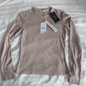 Ribbad ljusbrun långärmad tröja NA-KD - Säljer en ribbad långärmad tröja från NA-KD i en snygg ljusbrun nyans. Tröjan har rund hals och är tillverkad av återvunnen bomull, vilket gör den både skön och miljövänlig. Perfekt att styla med jeans eller kjol för en chill look.