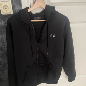 Svart hoodie från Polo Ralph Lauren - Svart hoodie från Polo Ralph Lauren med dragkedja, huva och snörning. Klassisk logga broderad i vitt på bröstet. Tröjan har långa ärmar och två fickor framtill. Perfekt för dig som gillar stilren streetwear och vill ha något bekvämt och snyggt. Har ej använt och den är helt ny.