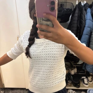 Vit stickad tröja från 157, S - Säljer en vit stickad tröja från 157 i storlek S. Tröjan har ett snyggt hålmönster och ribbade muddar vid hals, ärmslut och nederkant. Den är kortärmad och har en relaxed passform som är perfekt för vår och sommar. Superenkel att matcha med jeans eller kjol!