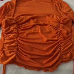 Orange omlott topp med snörning - Snygg orange topp med omlott-design och coola snörningsdetaljer på sidorna. Materialet är mjukt och glansigt, vilket ger en skön känsla mot huden. Perfekt för dig som vill sticka ut med färg och stil. Passformen är tight och toppen har långa ärmar.