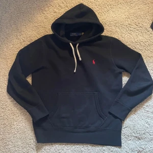 Marinblå hoodie från Polo Ralph Lauren - Marinblå hoodie från Polo Ralph Lauren med klassisk röd logobrodyr på bröstet. Tröjan har huva med vita snören, stor magficka och långa ärmar. Perfekt för chill dagar och enkel att matcha med jeans eller joggers.