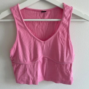 Rosa ribbad croppad linne - Säljer en karamellrosa croppat linne i storlek S. Toppen har ribbad struktur,  ärmlös. Perfekt för dig som gillar en enkel men snygg look med lite edge. Materialet är mjukt och stretchigt, vilket gör den superbekväm.