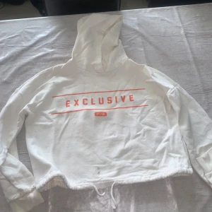 Vit hoodie med EXCLUSIVE-tryck - Vit hoodie med rött EXCLUSIVE-tryck på bröstet och snörning i nederkant. Croppad modell med huva och långa ärmar. Mjuk bomullskänsla, perfekt för chill dagar. Snygg streetstil som sticker ut med sin enkla design och coola tryck.