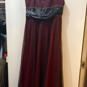 Vinröd långklänning med svart midja - Säljer en elegant vinröd långklänning med svart omlottband i midjan och små silvriga pärlor längs överdelen. Klänningen är axelbandslös och har ett skirt, flytande tyg som ger en snygg siluett. Perfekt för bal eller fest!
