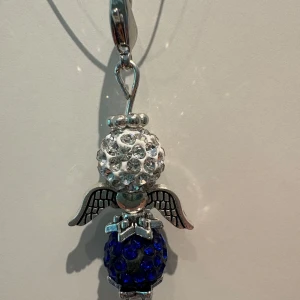 Halsband med ängel och strass - Unikt halsband med en ängelberlock i silverfärgad metall. Berlocken har en vit glittrig strassboll som huvud och en blå glittrig strassboll som kropp, samt detaljerade vingar och en liten rosett. Perfekt för dig som gillar personliga smycken med lite extra bling.