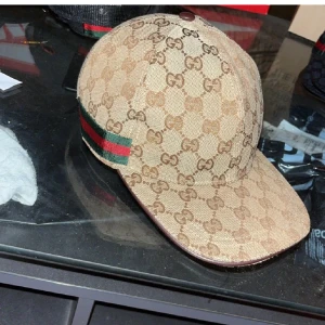 Gucci keps med GG-mönster, strl M - Exklusiv Gucci keps i beige med klassiskt GG-monogram över hela kepsen. Sidorna har gröna och röda ränder som ger den ikoniska Gucci-looken. Justerbar rem baktill i brunt läder. Tillverkad i bomullscanvas med brunt innerfoder.