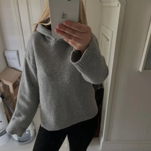 Grå stickad hoodie med huva - Mysig grå stickad hoodie med huva och långa ärmar. Tröjan har en loose passform och ribbade muddar vid ärmslut och nederkant. Perfekt för kyliga dagar. I bra skick därav att den är använd fåtal gånger.