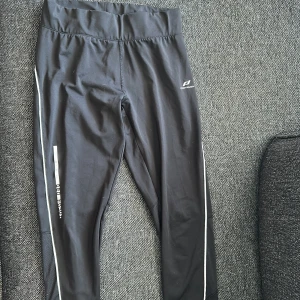 Svarta träningstights från Pro Touch - Svarta träningstights från Pro Touch med reflekterande detaljer längs benen och dragkedjor vid bensluten. Tightsen har hög midja, meshpaneler för extra ventilation och en liten ficka med dragkedja bak. Perfekta för löpning eller gymmet.