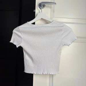 Vit Gina Tricot topp - Säljer en vit ribbad croppad topp med korta ärmar och vågig kant nertill.