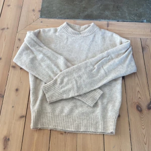 Beige stickad tröja med ribbad krage - Mysig beige stickad tröja från Cos i storlek 146/152. Tröjan har en ribbad hög krage och ribbade muddar vid ärmslut och nederkant. Perfekt för kyliga dagar och enkel att matcha med jeans eller mjukisbyxor. Lite nopprig på framsidan.