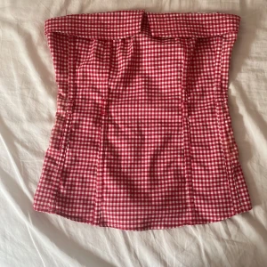 Rutig röd korsett från Ann Christine - Supersöt röd och vit rutig korsett från Ann Christine. Tubmodell med smockad rygg för extra stretch och dragkedja i sidan. Perfekt för dig som vill sticka ut med en retrovibe! 