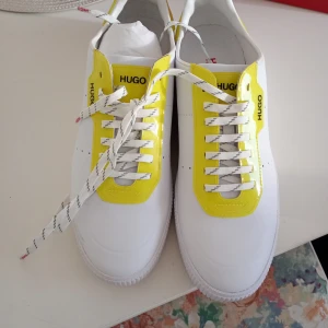 Vita och gula sneakers från Hugo - Fräscha sneakers från Hugo i vitt med glansiga gula detaljer runt snörningen. Skorna har vita snören med svarta ränder och en klassisk rund tå. Perfekta för dig som vill sticka ut med en färgklick i din outfit.