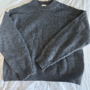 Grå stickad tröja från H&M - Mysig grå stickad tröja från H&M i storlek XS. Tröjan har en enkel design med rund hals och långa ärmar, perfekt för att hålla dig varm under kyliga dagar. Materialet är mjukt och har en lite fluffig känsla, vilket gör den extra skön att bära.