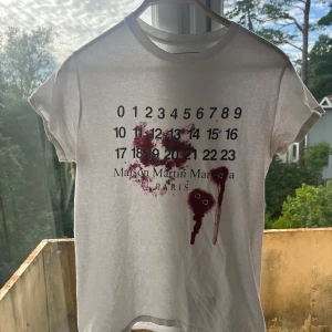 Maison Martin Margiela vit t-shirt med tryck - Säljer en vit t-shirt från Maison Martin Margiela med ikoniskt siffertryck och röd grafisk detalj framtill. T-shirten har rund hals och korta ärmar, tillverkad i mjuk bomull. Perfekt för dig som gillar statement-plagg och designer vibes.