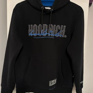Svart hoodie från Hoodrich med blå detaljer - Svart hoodie från Hoodrich med logga i grått och blått framtill. Klassisk känguruficka, huva med snörning och tryck även på ryggen. Mjuk insida och coola blå detaljer. Perfekt för dig som gillar streetwear och vill sticka ut. Den har även en liten ficka under armen som kan vara väldigt praktisk. Den är i storlek M men känns mer som en S. 