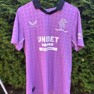 Lila Rangers FC fotbollströja Castore - Säljer en lila Rangers FC fotbollströja från Castore med svart krage och vit logga. Tröjan har geometriskt mönster, tryckta sponsorer som UNIBET och SEKO samt klubbmärke på bröstet. Tillverkad i lätt och ventilerande material, perfekt för match eller träning. Sällsynt tröja för dig som samlar på fotbollströjor. Har ni några funderingar är det bara att höra av sig! Priset är ej hugget i sten👍