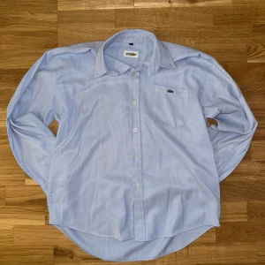 Ljusblå skjorta från Lacoste XL - 👔 Märke: Lacoste 🎨 Färg: Ljusblå  📏 Storlek: XL  📐 Mått: 	•	Bredd: 55 cm 	•	Längd: 70 cm  🧵 Material: Troligtvis bomull (lapp saknas)  ♻️ Skick: Bra skick! Inga hål eller fläckar. Använd men fräsch och välvårdad.  Hör av dig vid frågor eller funderingar. 