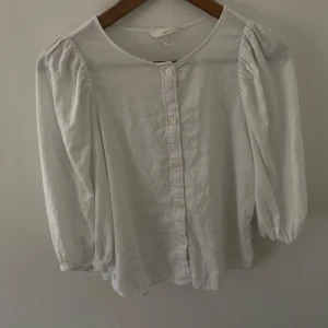 Vit blus med puffärm  - Snygg vit blus i luftigt linnematerial. Modellen har puffiga långärmar, rund halsringning och knäppning framtill med vita knappar. Perfekt för en stilren och trendig look.
