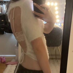 Vit fluffig topp med öppen rygg - Superfin vit topp i fluffigt material med korta ärmar och öppen rygg. Toppen har snörning i ryggen som ger en snygg detalj och croppad passform. Perfekt för dig som vill sticka ut med en unik och trendig look. Liten i storlek!