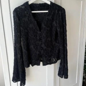 Svart blus med knappar - Säljer en svart blus med snyggt texturerat mönster. Blusen har v-ringning, långa ärmar och knappar framtill. Materialet är lätt. Använd ett fåtal gånger!💛💛