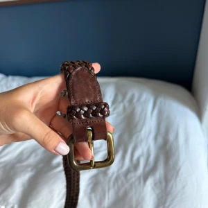 Flätat brunt skärp i skinn - Säljer ett snyggt, flätat bälte i mörkbrunt skinn med ett mässingfärgat metallspänne.