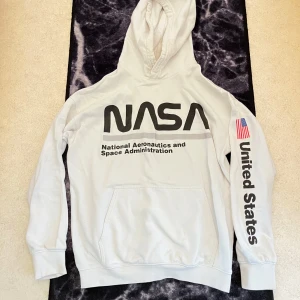 Vit NASA hoodie från H&M - Passar xxs-s.                                                            Säljer en vit hoodie från H&M med stor NASA-logga på bröstet och texten 'United States' samt amerikansk flagga på ena ärmen. Tröjan har huva, magficka och långa ärmar. Perfekt för dig som gillar rymden och vill ha en chill streetstyle.