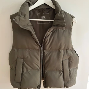 Croppad dunväst  - Snygg olivgrön dunväst från Zara med hög krage och dragkedja framtill. Västen har två stora fickor och en quiltad look. Perfekt att slänga över en hoodie eller tröja för extra värme.