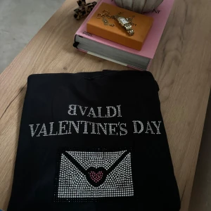 Svart Bvaldi Valentine's Day t-shirt - Svart t-shirt från Bvaldi med glittrande text 'VALENTINE'S DAY' och en stor strassdekorerad brev-ikon med hjärta framtill. Klassisk rund hals och korta ärmar. Perfekt statement-plagg med cool vibe och snygga detaljer. Storlek s