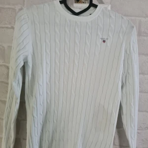 Vit kabelstickad tröja från GANT - Säljer en vit kabelstickad tröja från GANT med rund hals och broderad logga på bröstet. Tröjan är gjord i stretchig bomull och har långa ärmar. Perfekt för dig som gillar klassisk och clean stil med snygga detaljer.