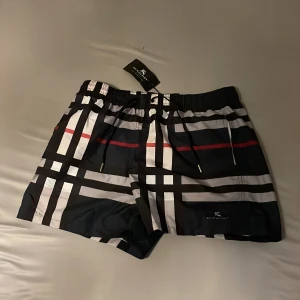 Burberry rutiga badshorts med snörning - Snygga badshorts från Burberry med klassiskt rutigt mönster i svart, vitt, grått och en röd linje. Shortsen har elastisk midja med snörning och är tillverkade i ett lätt syntetmaterial som torkar snabbt. Perfekt för stranden eller poolen.