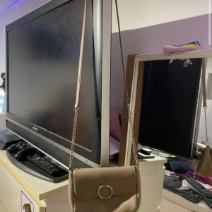 Beige axelväska med kedjerem - Säljer en stilren beige axelväska med silverfärgad kedjerem och snygg rund metallring på locket. Väskan har en kompakt och smidig form, perfekt för att bära det viktigaste. Passar till många olika outfits och är lätt att matcha.