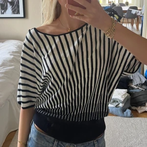 Randig vit och svart stickad tröja - Snygg stickad tröja med svart och vit randigt mönster, bredare midjemudd, korta ärmar och i såå skönt material. Tröjan har en cool detalj med svarta knappar längs ena axeln och en avslappnad, loose passform. 