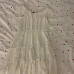 Vit volangklänning från KappAhl - Superfin vit klänning från KappAhl med flera lager volanger och en bred, rynkad krage. Klänningen är ärmlös och har en luftig, romantisk look med lätt transparent tyg. Perfekt för dig som gillar en söt och fräsch stil.