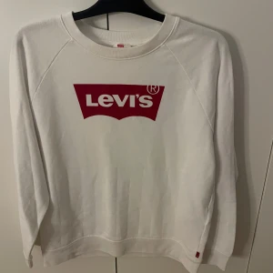 Vit sweatshirt från Levi's med röd logga - Vit sweatshirt från Levi's i storlek S med den klassiska röda Levi's-loggan tryckt på bröstet. Tröjan har rund hals och långa ärmar, tillverkad i mjuk bomull som är skön att ha på sig. Perfekt för dig som gillar enkel och stilren streetwear.