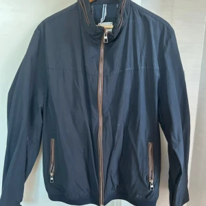 Svart vindjacka med dragkedjor - Snygg svart vindjacka med bruna detaljer längs dragkedjorna. Jackan har hög krage, två fickor med dragkedja och är tillverkad i lätt syntetmaterial. Perfekt för blåsiga dagar och enkel att matcha med det mesta. Långärmad och stilren design.