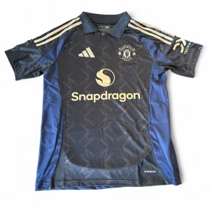 Manchester United blå/svart fotbollströja - Snygg Manchester United fotbollströja från Adidas med guldiga detaljer och loggor. Tröjan är svart med marinblå paneler och har subtilt geometriskt mönster. Kortärmad med klassisk krage och tre guldiga ränder på axlarna. Materialet är lätt och andas, perfekt för träning.