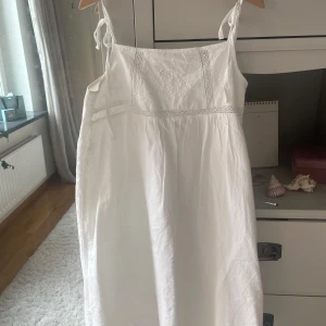 Vit sommarklänning med spetsdetaljer - Säljer en vit klänning i XXS med tunna axelband som knyts och vacker broderad spets upptill. Klänningen har en luftig passform och är tillverkad i mjukt bomullstyg, perfekt för varma dagar. Stilren och romantisk look med fina detaljer.