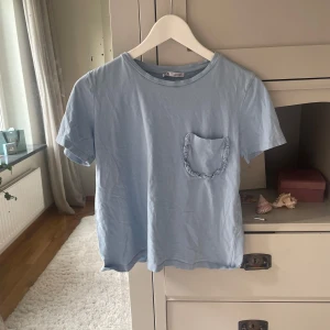 Ljusblå t-shirt med volangficka Zara - Snygg ljusblå t-shirt från Zara med korta ärmar och rund hals. På bröstet finns en liten ficka med volangkant som ger en gullig touch. Tillverkad i mjuk bomull som känns skön mot huden. Perfekt för dig som gillar enkel stil med en twist.