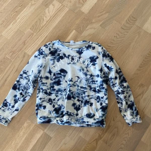 Blå tie-dye sweatshirt från Lindex - Säljer en oversized sweatshirt från Lindex med coolt blått och vitt tie-dye mönster och texten CALIFORNIA framtill. Tröjan har rund hals och långa ärmar, perfekt för en avslappnad och trendig look. Tillverkad i mjuk bomull.